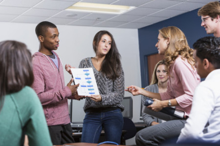 Multi-ethnic group of high school or university students and female teacher at school in classroom. Two students standing up, giving presentation to class. Main focus on the students holding the chart.
